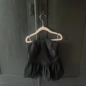 Svart volang tubtopp - Säljer en svart axelbandslös tubtopp med volang nertill. Toppen har en bandeauformad överdel och är perfekt för dig som vill ha en stilren men ändå lekfull look. Passar till både jeans och kjol för en trendig outfit.