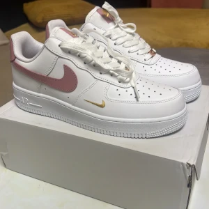Nike Air Force 1 vit/rosa/guld - Nike Air Force 1 sneakers i vitt skinn med rosa swoosh och detaljer i guld. Klassisk låg modell med perforerad tå, vita snören och rosa innerfoder. Snygg guldfärgad mini-swoosh på sidan och guldiga detaljer på plösen och snörningen. Pris kan diskuteras vid snabb affär!
