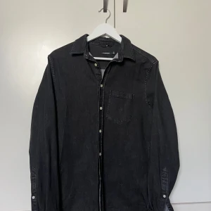 Svart jeansskjorta från J.Lindeberg - Svart jeansskjorta från J.Lindeberg i storlek M. Klassisk modell med bröstficka, krage och knappar framtill samt vid ärmslut. Skjortan har lång ärm och är tillverkad i ett slitstarkt jeanstyg som ger en cool och avslappnad vibe.