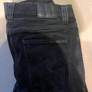 Svarta jeans från Nudie Jeans, W33 L34 - Säljer ett par svarta jeans från Nudie Jeans i klassisk femficksmodell. Jeansen har raka ben, snygga detaljer på bakfickan och logga på läderpatchen. Tillverkade i slitstarkt bomullstyg och passar perfekt till en avslappnad stil.