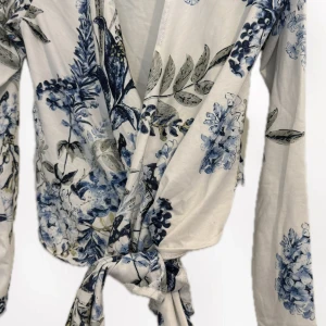 Blommig omlottopp med knytband - Supersöt omlottopp med blommigt mönster i blått och grönt på vit botten. Toppen har långa ärmar och knytband framtill som ger en snygg och avslappnad look. Perfekt för dig som gillar romantiska vibes och vill sticka ut lite extra.