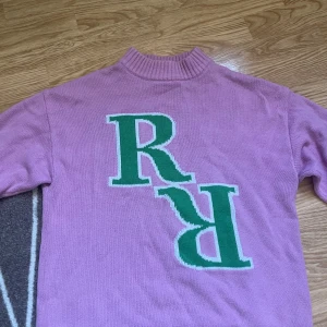 Rosa stickad tröja Gina Tricot - Rosa stickad tröja från Gina Tricot med högt ribbat krage och stora gröna bokstäver 'R' framtill. Tröjan har långa ärmar och ribbade muddar vid ärmslut och nederkant. Perfekt för dig som gillar färg och statement-detaljer.