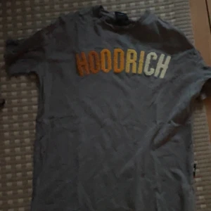 Grå HOODRICH t-shirt XS - Säljer en grå t-shirt från HOODRICH i storlek XS. T-shirten har korta ärmar och stor broderad text i gult och vitt framtill. Skön passform och perfekt för en avslappnad streetstil.