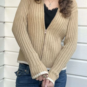 Beige ribbad kofta från Ralph Lauren - Ribbstickad beige kofta från Polo Jeans Co. Ralph Lauren med dragkedja framtill och vita detaljer vid ärmslut och nederkant. Tillverkad i 100% bomull och har långärmad passform. Perfekt för lager-på-lager och snygg till jeans.