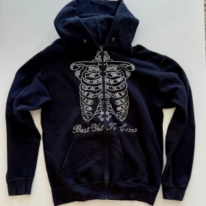 Svart hoodie med strass och skelettmotiv - Cool svart hoodie med huva och dragkedja framtill. Framsidan har ett stort skelettmotiv i strass och texten 'Best Yet To Come' under. Tröjan har känguruficka och långa ärmar, perfekt för dig som gillar edgy streetstyle.