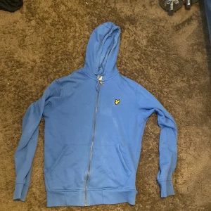 Blå hoodie från Lyle & Scott XS - Blå hoodie från Lyle & Scott i ekologisk bomull. Tröjan har huva, dragkedja framtill och två fickor. På bröstet sitter den klassiska gula fågel-loggan. Perfekt för chill dagar eller när du vill ha en enkel och snygg look.