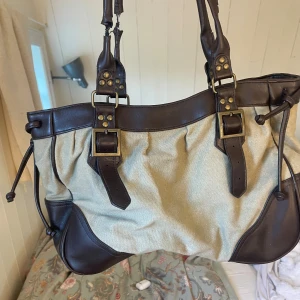 Beige och brun axelväska med spännen - Stilren axelväska i beige canvas och mörkbrunt skinn med snygga detaljer som spännen i mässing och knytband på sidorna. Väskan har rejäla handtag och coola kontraster mellan material och färger. Perfekt för dig som gillar en klassisk men edgy look. Skada i ena bandet!