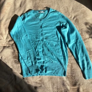 Turkos kofta från Gina Tricot - Snygg turkos kofta från Gina Tricot med rund halsringning och knappar hela vägen fram. Långärmad modell med ribbade muddar vid ärmslut och nederkant. Perfekt att slänga över en topp för en fräsch look.