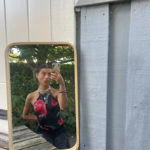 Svart och röd halterneck topp med jordgubbar - Unik halterneck topp i svart med stora röda jordgubbar och vit text. Toppen är ärmlös, har öppen rygg och knyts i nacken. Perfekt för dig som vill sticka ut med ett coolt och lekfullt plagg, eller styla med konstymbyxor. Y2K vibes.