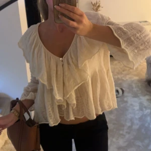 Vit blus med volang och v-ringning - Supersöt vitkrämig blus med v-ringning och stora volanger framtill. Blusen har trekvartsärmar med lätt utsvängd form och är i ett tunt, stickat material. Aldrig använd.