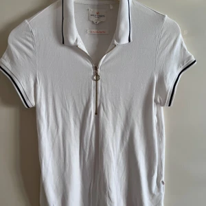 Vit piké-tshirt Holly & Whyte - Vit piké-tshirt med svart kant på krage och ärmslut. Dragkedja framtill med ringdetalj. Klassisk och sportig look, perfekt till jeans. Mjukt bomullsmaterial som känns skönt mot huden.