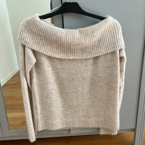 Beige offshoulder stickad tröja Gina Tricot - Mysig beige stickad tröja från Gina Tricot med bred ribbad offshoulder-krage. Tröjan har lång ärm och är perfekt för kyliga dagar. Den är mjuk och har en avslappnad passform som är enkel att styla med jeans eller kjol.