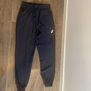 Svarta träningsbyxor från Asics - Säljer ett par svarta träningsbyxor från Asics med elastisk midja och dragsko. Byxorna har smal passform vid anklarna och är tillverkade i ett lätt syntetmaterial. Perfekta för träning eller chill. Asics-logga på låret.