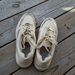 Beige sneakers med gulddetaljer - Snygga beige sneakers med guldfärgade detaljer och perforerade partier på ovandelen. Skorna har snörning och en brun sula med bra grepp. Tillverkade i syntetmaterial och passar perfekt för dig som gillar en clean men ändå trendig look.