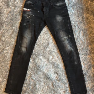 Svarta skinny jeans från Dsquared2 - Högra fickan är sönder men sytt igen så man får bara plats med airpods och nycklar ej mobil men de andra fickorna är som de ska. Svarta jeans från Dsquared2 med skinny passform och coola slitningar, färgstänk och patchdetaljer. Jeansen har fem fickor, röd läderpatch bak och unika detaljer framtill. Tillverkade i stretchig denim för en bekväm känsla.