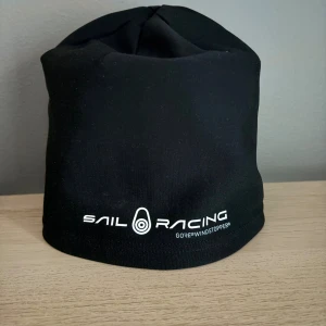Svart mössa Sail Racing Gore-Tex - Svart mössa från Sail Racing med vit logga framtill. Tillverkad i vindtätt Gore-Tex-material, perfekt för blåsiga dagar. Enkel och stilren design med diskret etikett på sidan. Sitter skönt och skyddar mot kyla.