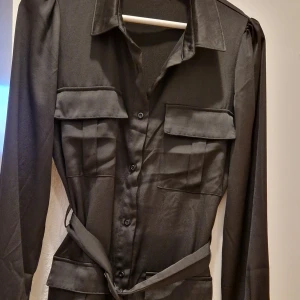 Svart långärmad skjortklänning Bik Bok - Snygg svart skjortklänning från Bik Bok i storlek S. Klänningen har långa ärmar med manschett, fyra stora fickor fram, knappar hela vägen och ett matchande bälte i midjan. Materialet är mjukt och har en lätt glansig finish som ger en cool look.