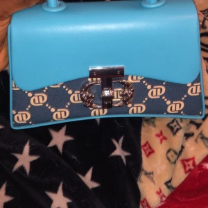 Blå axelväska med monogram och silverdetaljer - Snygg axelväska i ljusblått med avtagbar axelrem och handtag. Framsidan har blått tyg med vitt monogrammönster och coolt silverlås framtill. Väskan är tillverkad i syntetmaterial och har en stilren, modern look som sticker ut.