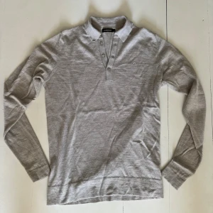 Grå pikétröja från J.Lindeberg - Snygg grå pikétröja från J.Lindeberg med klassisk krage och tre knappar framtill. Tröjan är långärmad och har en diskret logga broderad på bröstet. Tillverkad i mjukt material som känns skönt mot huden.
