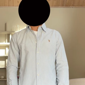 Randig skjorta från Ralph Lauren - Säljer en snygg blå- och vitrandig skjorta från Ralph Lauren i super slim fit. Skjortan har klassisk krage, knappar framtill och den ikoniska broderade loggan på bröstet. Tillverkad i mjuk bomull och har långa ärmar. Perfekt för en clean och stilren look. Är själv 173 passar perfekt 