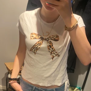 Vit t-shirt med leopardrosett - Vit t-shirt med korta ärmar och ett stort tryck av en rosett i leopardmönster på bröstet. Modellen är figurnära och har en klassisk rund halsringning. Perfekt för dig som vill sticka ut med en cool detalj.