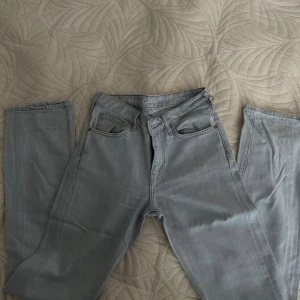 Ljusgrå raka jeans lågmidjade - Säljer ett par ljusgrå jeans med rak passform och  lowwaist. Perfekta för en clean och avslappnad look. Jeansen är i ett mjukt denimtyg och har diskreta detaljer i silver och koppar.