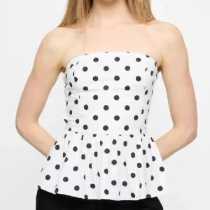 Prickig vit tubtopp - Snygg bandeau tubtopp i vit med svarta prickar och volang nedtill. Toppen är axelbandslös och har en feminin, lätt utsvängd form som ger en cool och trendig vibe. Perfekt för dig som gillar att sticka ut med mönster och vill ha en fräsch look. Ser lite längre ut i verkligheten