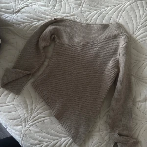 Beige ribbstickad tröja med asymmetrisk kant - Säljer en beige ribbstickad tröja med lång ärm och snygg asymmetrisk nederkant. Tröjan har en bredare halsringning och är gjord i mjukt stickat material, perfekt för lager-på-lager och chill vibes.