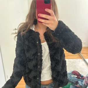 Svart pälsjacka med knappar - Mysig svart pälsjacka med knäppning framtill. Jackan har en fluffig och mjuk känsla, perfekt för att hålla värmen. Långärmad och rak passform som funkar till många olika outfits. Snygg och enkel att styla till både jeans och klänning.
