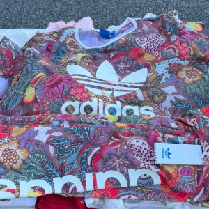 Adidas set i färg - Färgstarkt set från Adidas med stort vitt logotryck längs benet och T-shirt. Mönstret är blommigt i rosa, rött, gult, grönt och blått. Materialet är stretchigt och mjukt, perfekt för dig som gillar att sticka ut. Snygga till sneakers och hoodie.