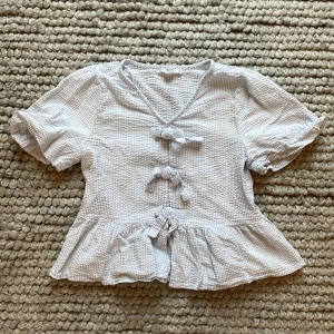  Blå vit Randig blus med rosetter från Gina Tricot nytt skick men tvättad ett par gånger - Supersöt blus från Gina Tricot i ljusblått och vitt med randigt mönster. Blusen har v-ringning, tre knyt framtill och volang nertill. Puffiga korta ärmar och mjukt, strukturerat bomullstyg gör den perfekt för vår och sommar.