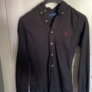 Svart skjorta från Ralph Lauren slim fit - Svart långärmad skjorta från Ralph Lauren i slim fit-modell. Skjortan har klassisk krage, vita knappar och rött broderat Polo-logo på bröstet. Tillverkad i bomull och passar perfekt för en clean och stilren look. Perfekt nu inför hösten