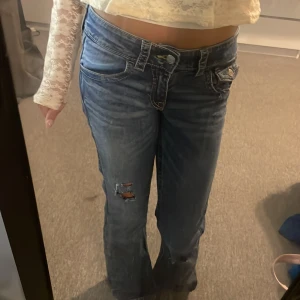 Blå bootcut jeans med slitningar - Säljer ett par blå bootcut jeans med låg midja och slitningar på benen för en cool, avslappnad vibe. Jeansen har klassiska fickor med knappdetaljer och är tillverkade i denim. Endast använd en gång💞 pris går såklart att diskutera💞💞