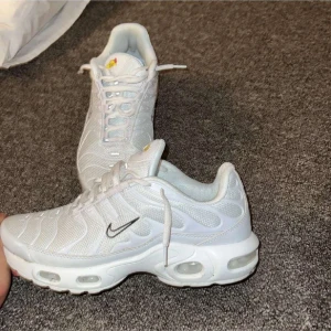 Vita Nike Air Max Plus sneakers - Säljer ett par fräscha vita Nike Air Max Plus sneakers med mesh- och syntetöverdel, ikoniska vågiga detaljer och synliga Air-enheter i sulan. Skorna har svart Swoosh-logga på sidan och klassisk snörning. Perfekta för dig som gillar sportig och clean stil.