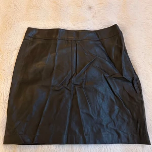 Svart skinnkjol från Gina Tricot - Snygg svart kort skinnkjol från Gina Tricot i storlek 36. Kjolen har en stilren design med dragkedja i sidan och markerad midja. Perfekt för dig som vill ha en edgy och trendig look.