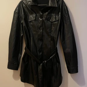 Svart skinnjacka med bälte, stl S - Snygg svart skinnjacka i längre modell med bälte i midjan och stora knappar framtill. Jackan har klassisk krage, två bröstfickor med lock och långa ärmar. Perfekt för dig som vill ha en edgy och stilren look.