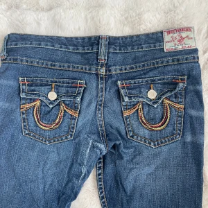 True Religion blå bootcut jeans - Snygga blå jeans från True Religion med ikoniska broderade bakfickor i rött, gult och orange. Klassisk bootcut-modell med kontrastsömmar och silverfärgade knappar. Jeansen har dragkedja och är tillverkade i slitstarkt denimtyg. Har behöft sy där bak men inget som syns när man har på sig de