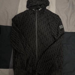 Svart vindjacka från Dior med monogram - Snygg svart vindjacka från Dior med ikoniskt monogrammönster över hela jackan. Jackan har huva, dragkedja framtill och en metallbadge på ärmen. Perfekt för dig som vill sticka ut med en exklusiv streetstyle-look.