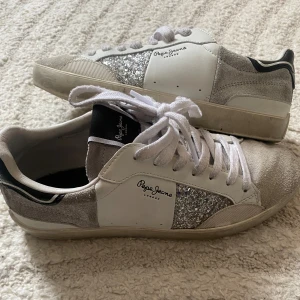 Glittriga sneakers från Pepe Jeans - Snygga sneakers från Pepe Jeans i vitt och grått med glittriga silverdetaljer på sidorna. Skorna har snörning, rund tå och en platt sula. Ovandelen är i en mix av skinn och mocka med coola kontraster och svart detalj på hälen. Ganska använda, men väldigt bra skick. Pris går att diskuteras!