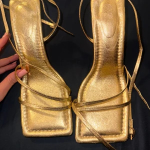 Guldiga sandaler med smala remmar - Snygga sandaler i metallic guld med smala remmar som snörs runt foten och vristen. Skorna har fyrkantig tå, hög klack och platt sula framtill. Perfekta för att sticka ut med en glittrig touch. Tillverkade i syntetmaterial.