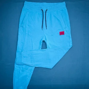 Blå mjukisbyxor från Hugo - Ljusblå mjukisbyxor från Hugo med svart snörning i midjan och ribbade muddar vid bensluten. Byxorna har två sidofickor, en bakficka och en röd Hugo-logga framtill. Sköna och stilrena, perfekta för chill dagar. Priset kan diskuteras!!!