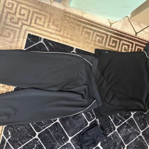 Svarta träningsbyxor och T-shirt från deVal S - Svarta korta träningsbyxor och T-shirt från deVal i storlek S. Även en simnings mössa från SOC. Byxorna och T-shirt har en sportig look med smal passform, elastisk midja och diskreta vita detaljer längs sidorna. Tillverkade i mjukt och stretchigt syntetmaterial som andas, perfekta för träning, simning eller chill. Byxorna har en liten inre ficka som är för skåp nyckeln