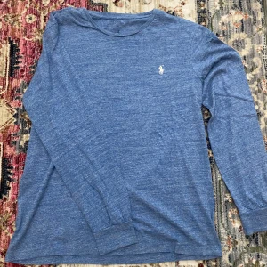 Blå långärmad tröja från Polo Ralph Lauren - Snygg blå långärmad tröja från Polo Ralph Lauren med klassisk rund hals och vit broderad logga på bröstet. Tröjan är i mjuk bomull och har ribbade muddar vid ärmsluten. Perfekt för en chill och stilren look.