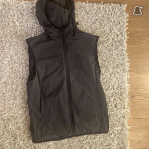 Svart dunväst från Massimo Dutti - Svart dunväst med huva från Massimo Dutti. Västen har en stilren look med dragkedja framtill och två sidofickor. Materialet är syntet och dun, perfekt för lager-på-lager. Passformen är normal och huvan har justerbara snören för extra funktion.