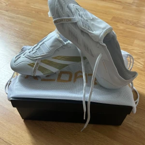 Vita och guldiga Adidas Predator - Säljer ett par stilrena Adidas Predator fotbollsskor i vitt med guldiga detaljer och snörning. Skorna har en modern design med klassiska tre ränder på sidan och levereras med tillhörande påse. Perfekta för dig som vill sticka ut på planen.