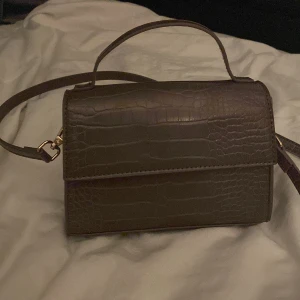 Brun axelväska i croco-imitation - Säljer en brun axelväska med croco-mönstrad yta och guldiga detaljer. Väskan har både handtag och lång axelrem, samt magnetlås och ett rymligt fack inuit.