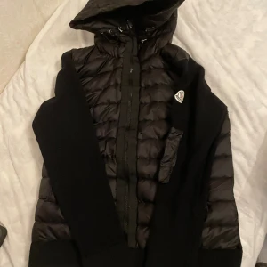Svart Moncler pufferjacka med huva - Säljer en svart pufferjacka från Moncler med stickade ärmar och quiltad kropp. Jackan har huva, dragkedja framtill och Moncler-logga på bröstet. Perfekt för dig som vill ha en stilren och varm jacka med snygga detaljer.