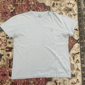 T-shirt Polo Ralph Lauren - Snygg ljusblå t-shirt från Polo Ralph Lauren med v-ringning och klassisk orange broderad logga på bröstet. T-shirten är kortärmad och gjord i mjuk bomull, perfekt för en clean och stilren look.