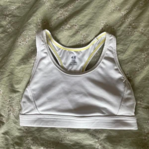 Vit sporttopp från H&M Sport S - Säljer en vit sporttopp från H&M Sport i storlek S. Toppen har brottarrygg, gula kontrastsömmar och ett litet gult logomärke på ryggen. Tillverkad i stretchigt syntetmaterial som passar perfekt för träning och aktiva dagar.