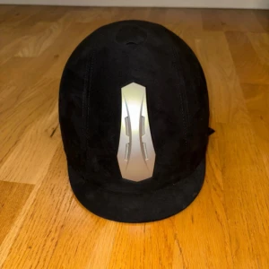 Svart ridhjälm CRW med silverdetalj - Snygg svart ridhjälm från CRW med en cool silverfärgad detalj framtill. Hjälmen har klassisk form, justerbart hakband och är klädd i mockaliknande material. Perfekt för dig som vill ha stil på ridbanan.
